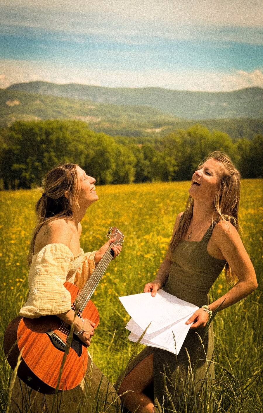 The Twin Heart Sound duo laughing in a summer meadow, one holding a guitar, the other holding lyric sheets